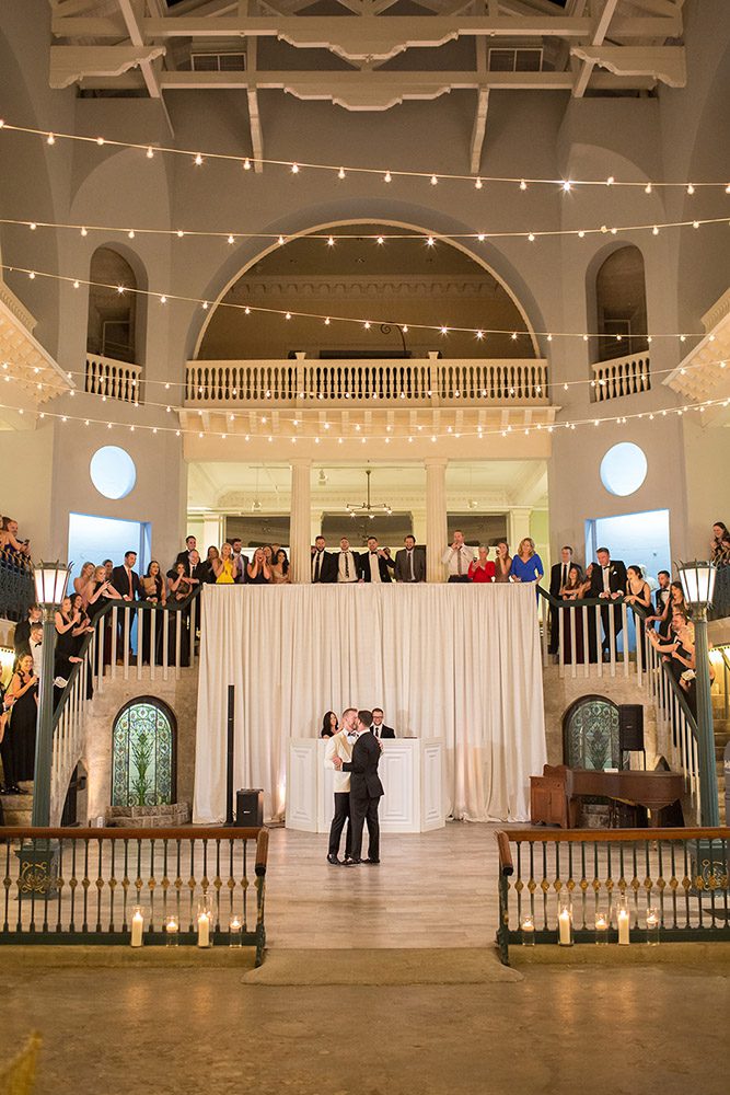 grooms sharing a first dance while their guests watch from the staircase