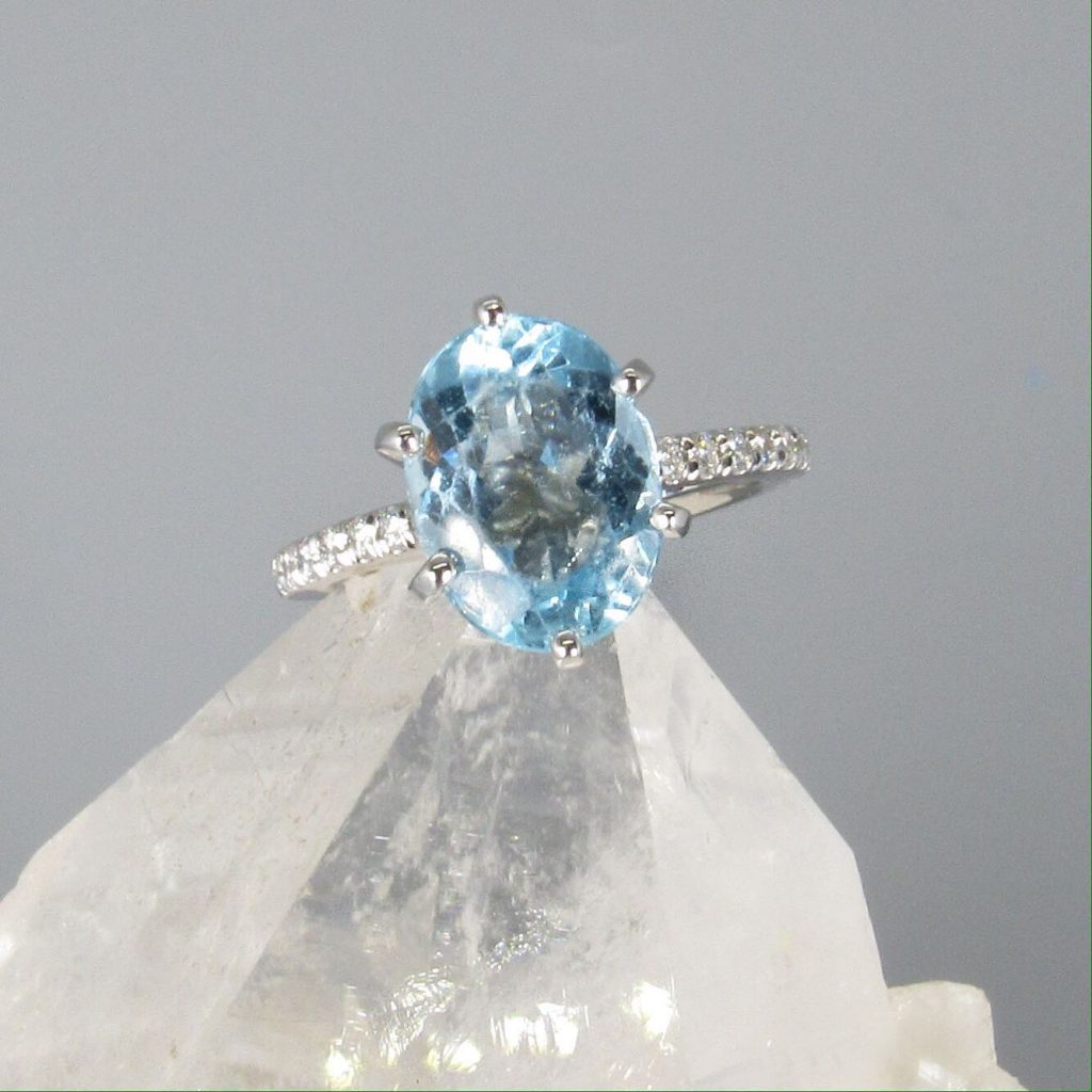 A blue, oval shaped diamond engagement ring with a diamond encrusted band.