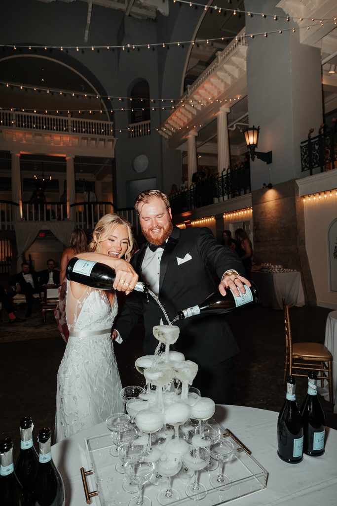 Newly married husband and wife pouring champagne into a large champagne tower during their wedding reception.