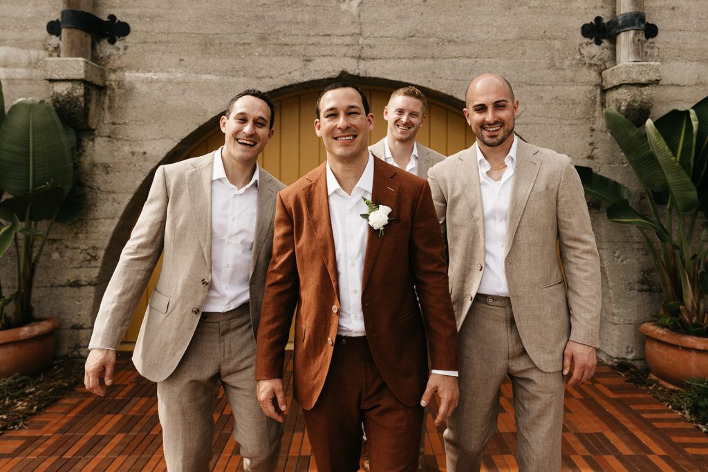 A groom wearing a rust colored suit for his wedding, surrounded by his groomsmen wearing taupe color suits. 