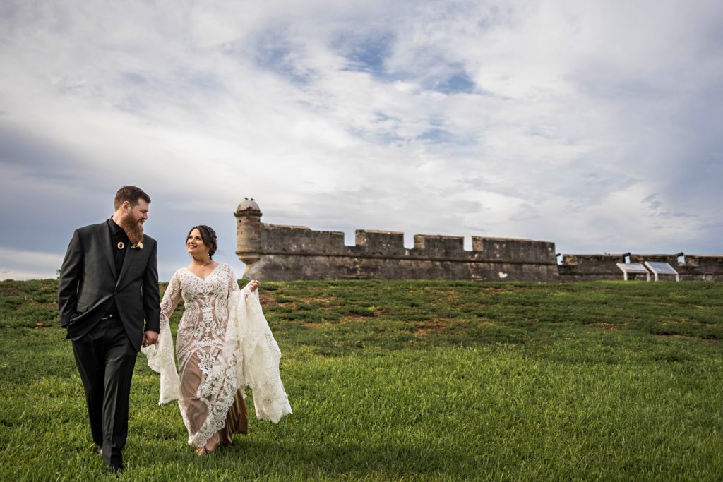 Bride and groom walking through field with Spanish fort in the background