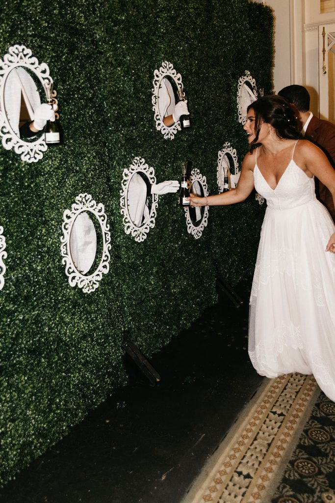 multiple hands reaching through greenery wall to offer champagne