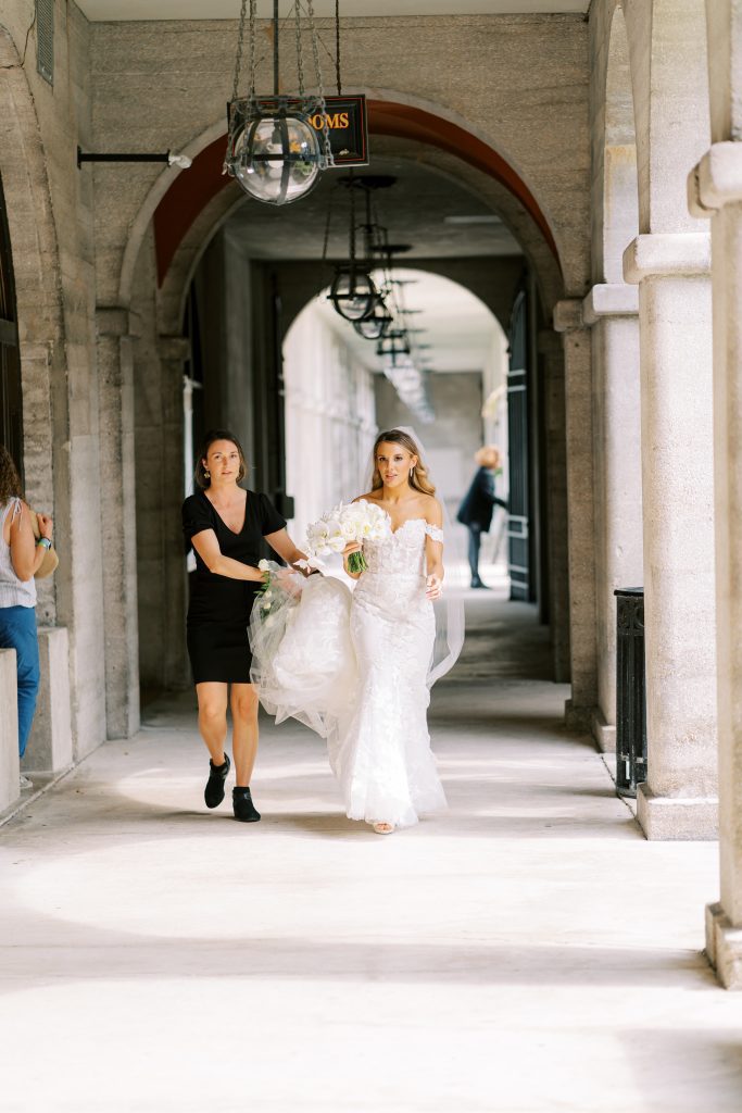 wedding planner holds bride's train as she walks through portico