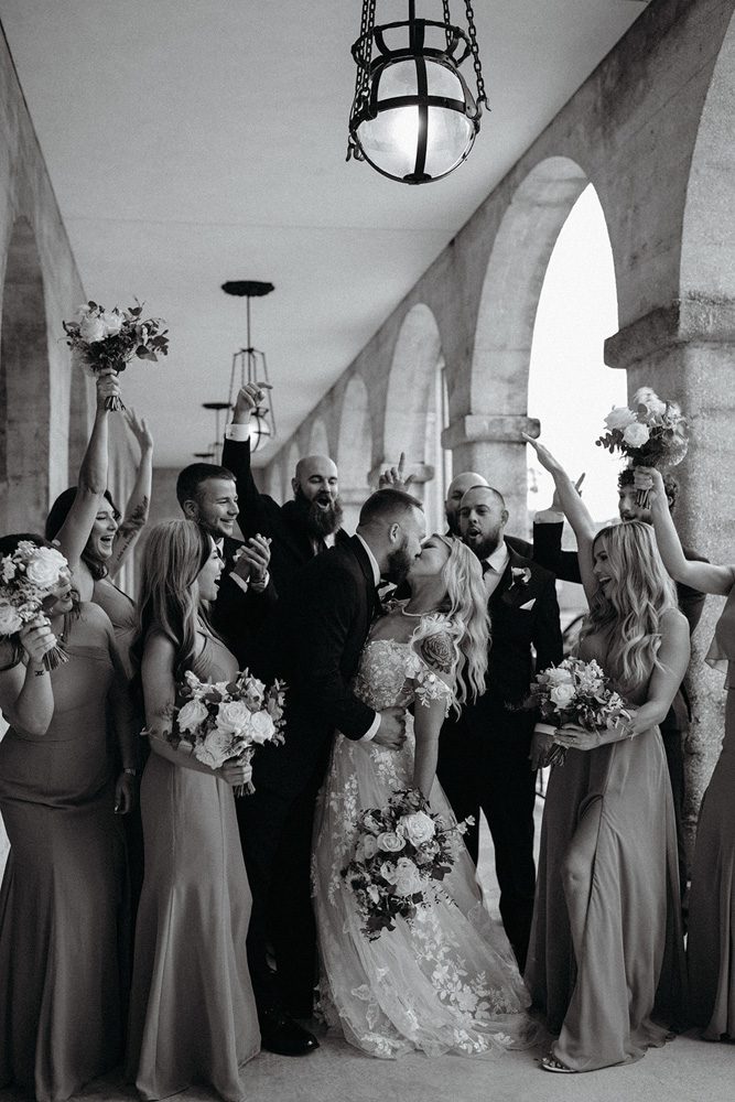 Black and white photo of bride and groom kissing as wedding party cheers
