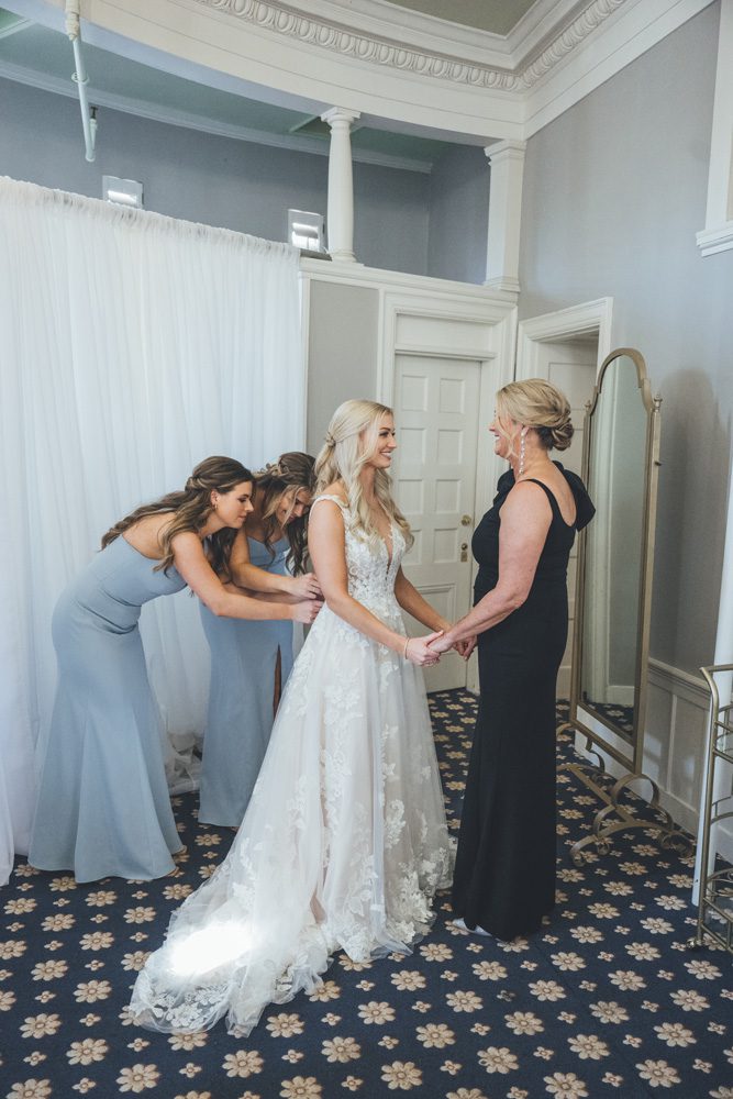 Bride and her mother while bridesmaids help zip her dress.